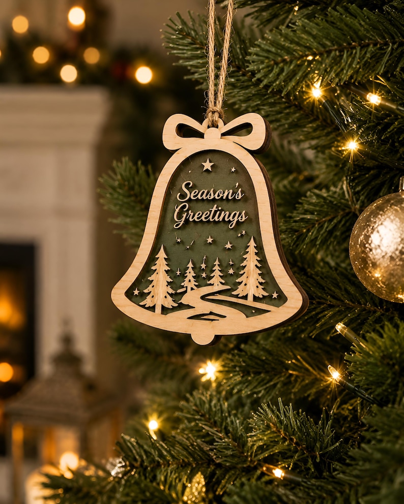 Op de afbeelding: Een klokvormige houten kerstornament met een gelaagd ontwerp en een donkergroene achtergrond. De uitsparing toont een kronkelend pad door een bos van dennenbomen onder een sterrenhemel, met de tekst Season's Greetings in een sierlijk lettertype.