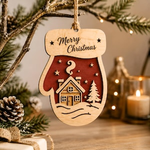 Op de afbeelding: Een houten kerstornament in de vorm van een want met een gelaagd ontwerp en een donkerrode achtergrond. De uitsparing toont een klein huisje met een schoorsteen en een dennenboom, geaccentueerd door sterren. De tekst Merry Christmas staat bovenaan.
