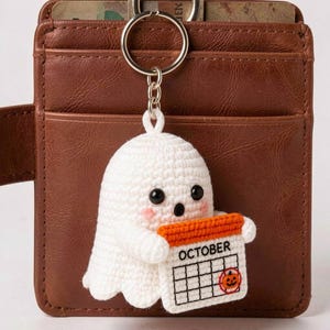 Puede incluir: Un llavero de fantasma tejido a ganchillo en color blanco con ojos de cuentas negras, mejillas rosadas y una boca pequeña. El fantasma sostiene una página de calendario rectangular blanca con borde naranja, el texto OCTOBER y una calabaza.