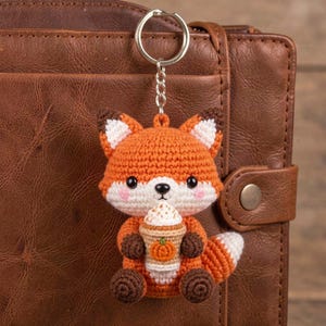 May include: A crocheted fox keychain featuring an orange and white fox holding a tan coffee cup with a small orange pumpkin icon on the front. The fox has black eyes, a black nose, and pink cheeks, attached to a silver metal split ring keychain.