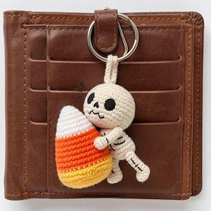 May include: A brown leather bi-fold wallet featuring a crochet keychain of a cream-colored skeleton holding a large, triangular candy corn piece with white, orange, and yellow horizontal stripes. The skeleton has black embroidered eyes and mouth.