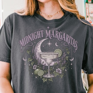 May include: A charcoal gray short-sleeve t-shirt featuring a graphic print of a margarita glass, a crescent moon, and stars. The text "MIDNIGHT MARGARITAS" is arched above the illustration, which is surrounded by purple flowers and green leaves.