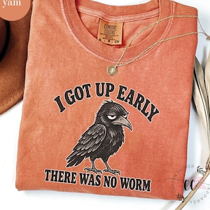 May include: A terracotta-colored crew neck t-shirt featuring a black graphic of a grumpy crow. The text above the crow reads I GOT UP EARLY and the text below reads THERE WAS NO WORM. The shirt has a Comfort Colors brand label at the neck.