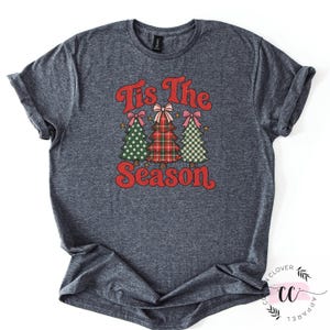 May include: A dark heather gray short-sleeve t-shirt featuring a graphic of three stylized Christmas trees with pink bows. The text "Tis The Season" is printed in red above and below the trees, which feature polka dot, plaid, and checkered patterns.