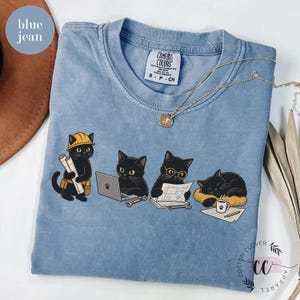 May include: A blue jean colored t-shirt featuring a graphic of four black cats performing various tasks: one wearing a yellow hard hat holding blueprints, one using a laptop, one wearing glasses reading plans, and one sleeping on a yellow bed.