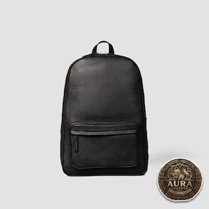Op de afbeelding: Een zwarte leren rugzak met een groot hoofdvak en een kleiner voorvak met ritssluiting. De tas heeft een handvat aan de bovenkant en verstelbare schouderbanden. Een cirkelvormig logobadge met de tekst Aura Leather is in de hoek zichtbaar.
