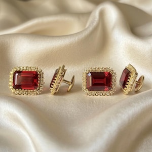 May include: Two pairs of rectangular cufflinks featuring a deep red, emerald-cut gemstone center surrounded by a halo of small, clear, sparkling stones set in a polished gold-tone metal frame with a standard T-bar swivel backing mechanism.