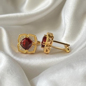 Puede incluir: Un par de gemelos dorados con una piedra preciosa roja oscura facetada de forma cuadrada en el centro. La piedra está rodeada por un borde de pequeñas piedras claras engastadas en pavé dentro de un marco dorado texturizado, con cierre giratorio.