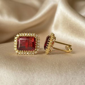 Puede incluir: Un par de gemelos dorados con una piedra preciosa rectangular de color rojo intenso en el centro. Cada piedra está rodeada por un halo de pequeñas piedras redondas transparentes engastadas en un marco dorado con cierre de barra en T.