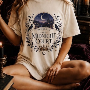以下が含まれることがあります： 「MEMBER OF THE MIDNIGHT COURT」という文字がプリントされたクリーム色の半袖Tシャツ。デザインには、三日月、城のシルエット、2羽の黒い鳥、葉のついた枝、そして下部には王冠が描かれています。