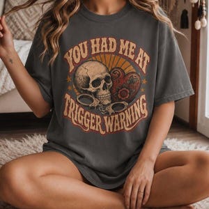 Könnte beinhalten: Ein anthrazitfarbenes Kurzarm-T-Shirt mit einem Grafikdruck, der einen menschlichen Schädel, ein Herz mit floralen Details und Handschellen zeigt. Der Text YOU HAD ME AT TRIGGER WARNING ist in einer geschwungenen Retro-Schrift gedruckt.