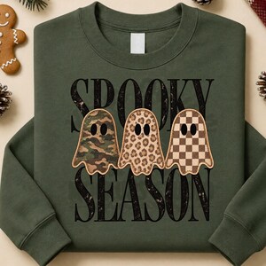 Op de afbeelding: Een olijfgroene sweater met ronde hals en de zwarte tekst SPOOKY SEASON. Onder de tekst staan drie spookillustraties met verschillende patronen: één met camouflageprint, één met luipaardprint en één met een bruin dambordpatroon.