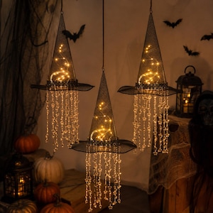 May include: Three hanging witch hats made of black wire mesh, each illuminated by internal warm white string lights. Each hat features dangling strands of clear star-shaped beads hanging from the brim, creating a glowing, layered effect.