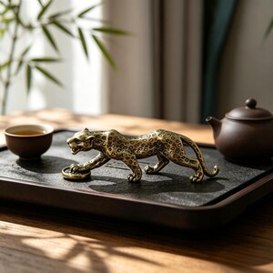 以下が含まれることがあります： 金色の金属製ヒョウの置物。忍び寄るような姿勢で、詳細な斑点と質感のある表面が特徴です。ヒョウは片足を小さな円形のコインのような物の上に乗せており、磨き上げられた仕上げが施されています。