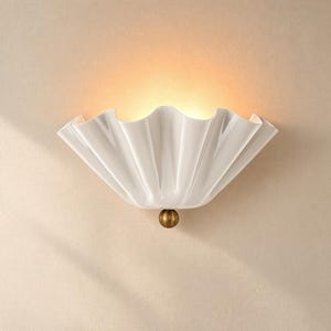 Può includere: Una lampada da parete bianca con un paralume plissettato a forma di ventaglio e bordo smerlato. L'apparecchio è illuminato dall'interno, emettendo una luce calda, ed è fissato alla base da un piccolo elemento decorativo sferico color ottone.