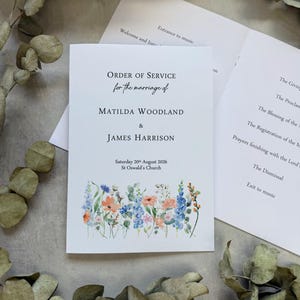 May include: A white paper wedding order of service booklet featuring black text that reads Order of Service for the marriage of Matilda Woodland & James Harrison, Saturday 20th August 2026, St Oswald's Church, with a colourful floral illustration.