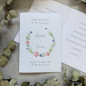 May include: A white wedding order of service booklet featuring a floral wreath illustration with pink roses and blue cornflowers. The text reads Order of Service for the marriage of Matilda & James, Saturday 20th August 2026, St Oswald's Church.