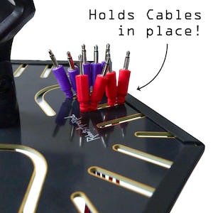 Puede incluir: Organizador de cables negro con detalles dorados, sosteniendo cables de audio rojos y morados. Se muestra el texto "Holds Cables in place!". El producto está etiquetado como "Robots Are Fed".