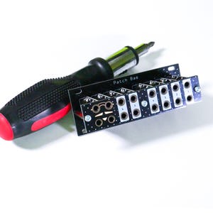 Pu&ograve; includere: Un cacciavite nero e rosso accanto a un circuito stampato nero con la scritta "Patch Bae". Il circuito stampato presenta diverse aperture circolari ed &egrave; progettato per componenti elettronici.