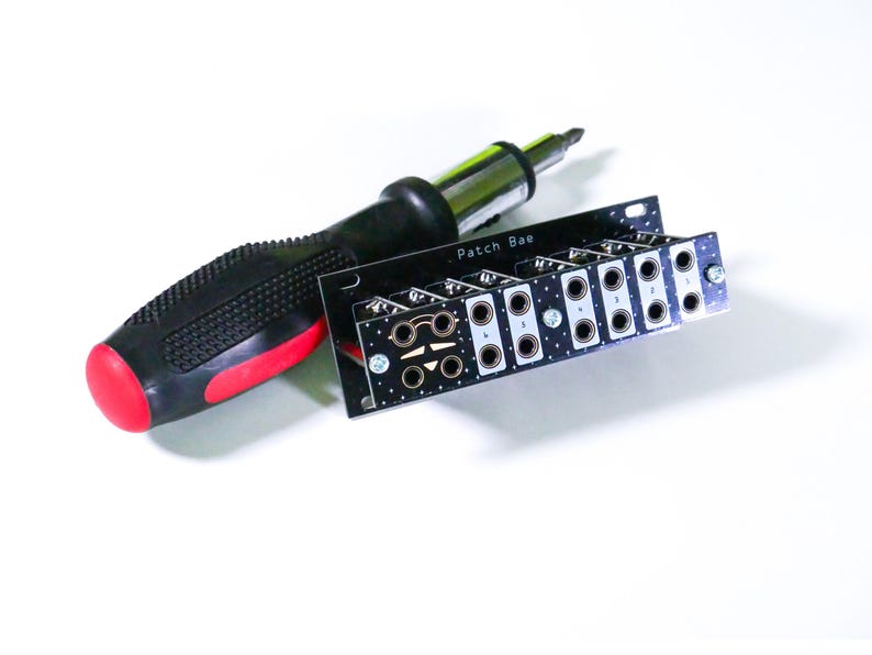 Pu&ograve; includere: Un cacciavite nero e rosso accanto a un circuito stampato nero con la scritta "Patch Bae". Il circuito stampato presenta diverse aperture circolari ed &egrave; progettato per componenti elettronici.