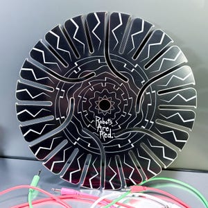 May include: A black and white circular art piece with a geometric design and the text "Robots Are Red." Several colorful audio cables are in the foreground. The art piece has a radial pattern with a central star-like shape.