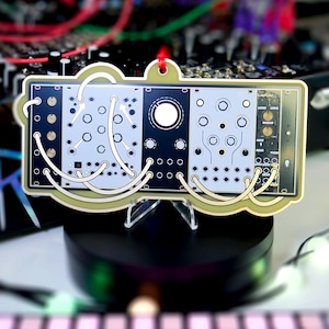 Peut inclure: Ornement décoratif en forme de panneau de synthétiseur modulaire. Le panneau est principalement noir et gris clair, avec de nombreux boutons circulaires et points de connexion. Des lignes blanches incurvées relient les composants. Bordure dorée et boucle rouge.