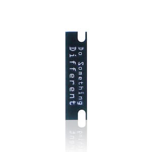 May include: A rectangular black tag with rounded corners and a small circular hole at the top and bottom. The tag features the white text "Do Something Different" printed vertically in a clean, sans-serif font centered on the black surface.