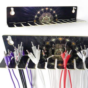 Puede incluir: Organizador de cables negro con un diseño floral decorativo. El organizador tiene múltiples ranuras para sujetar varios cables de audio. Cables de diferentes colores, incluyendo morado, rojo y blanco, están enchufados.