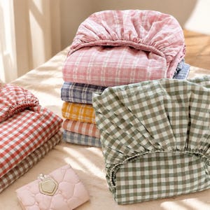 Op de afbeelding: Een stapel opgevouwen hoeslakens met diverse gingham ruitpatronen in roze, blauw, geel, oranje en groen. Een apart rood-wit gingham laken ligt opgevouwen aan de zijkant en een klein, lichtroze gewatteerd tasje ligt op de voorgrond.