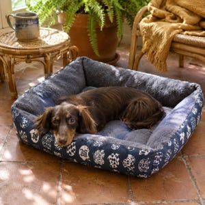 May include: A long-haired brown dachshund resting in a square pet bed. The bed features a dark blue exterior with a white repeating pattern of stylized animal shapes and a soft, plush, dark gray interior lining with raised side walls.