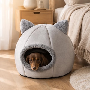 May include: A light gray, dome-shaped pet bed with two plush, textured cat-like ears on top. The bed features a circular opening with a fuzzy, dark gray interior lining. A brown dachshund is resting inside the enclosed, soft-sided structure.