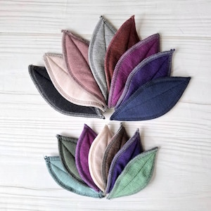 May include: An overhead shot displays two arrangements of leaf-shaped fabric items. The leaves are in various colors, including shades of blue, purple, green, and pink. Each leaf has a contrasting stitch around the edge, and they are arranged on a light wood-grain surface.