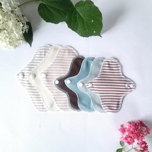 May include: Six reusable cloth menstrual pads in various colors and patterns, including white, brown, blue, and striped. The pads are arranged in a stack, with the white pad on top and the striped pad on the bottom.