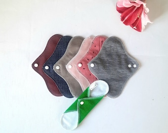 May include: A set of six reusable cloth menstrual pads in various colors and patterns, including red, pink, gray, and brown. The pads are folded and arranged in a stack. A green cloth pad is folded and placed in front of the stack.