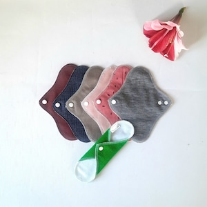 May include: A set of six reusable cloth menstrual pads in various colors and patterns, including red, pink, gray, and brown. The pads are folded and arranged in a stack. A green cloth pad is folded and placed in front of the stack.
