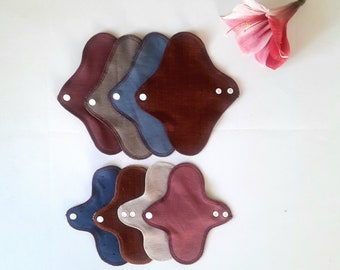 May include: A set of six reusable cloth menstrual pads in various shades of brown, blue, and beige. The pads are arranged in two stacks of three, with the top stack slightly offset from the bottom stack. Each pad has a snap closure and a soft, absorbent fabric.