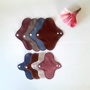 May include: A set of six reusable cloth menstrual pads in various shades of brown, blue, and beige. The pads are arranged in two stacks of three, with the top stack slightly offset from the bottom stack. Each pad has a snap closure and a soft, absorbent fabric.