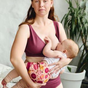 Op de afbeelding: Een vrouw in een bordeauxrode bh en bijpassende ondergoed is een baby aan het borstvoeden. De baby draagt een wit shirt en een luier met een patroon.