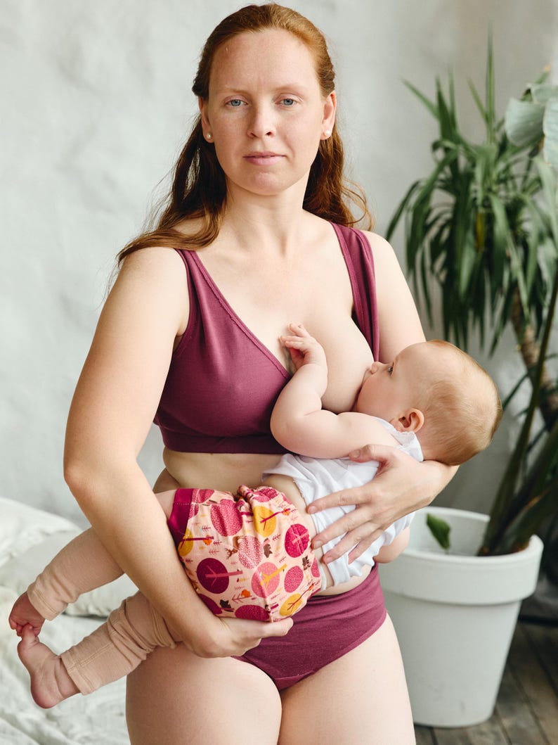 Op de afbeelding: Een vrouw in een bordeauxrode bh en bijpassende ondergoed is een baby aan het borstvoeden. De baby draagt een wit shirt en een luier met een patroon.