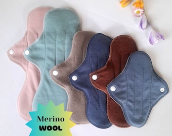 May include: A set of six reusable cloth menstrual pads made of merino wool in various colours, including pink, blue, brown, and grey. The pads have a starburst-shaped label that reads "Merino Wool".