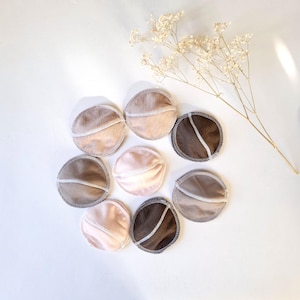 May include: Eight reusable breast pads in various shades of brown, beige, and pink. The pads are circular and have a sewn edge. They are arranged in a circle on a white surface.