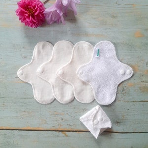 May include: Four white reusable cloth menstrual pads with a white cotton pad folded in the center. The pads are on a light blue wooden surface with a pink flower and purple flowers in the background.