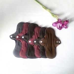 May include: Five reusable cloth menstrual pads in different colors, including brown, burgundy, and gray. The pads are arranged in a row, with the top pad being the darkest brown and the bottom pad being the lightest gray.