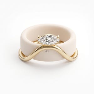 May include: A thick, matte beige silicone band paired with a polished gold-tone metal ring featuring a marquise-cut clear stone. The gold ring has a curved, wave-like design and includes the letters AD engraved on the front surface.