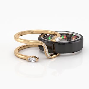 Puede incluir: Un anillo abierto de oro con un diamante de talla marquesa en cada extremo y una hilera de pequeños diamantes redondos a lo largo de la banda curva, colocado frente a un anillo ancho de cerámica negra con sensores electrónicos internos.
