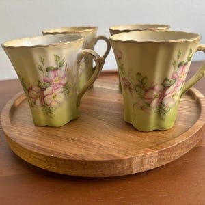 Puede incluir: Un juego de cuatro tazas de cerámica de color amarillo pálido con bordes festoneados y un patrón floral rosa pintado con hojas verdes. Cada taza tiene un asa curva y un borde acampanado, dispuestas sobre una bandeja redonda de madera.