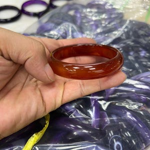 May include: A smooth, polished, translucent reddish-orange bangle bracelet with a rounded profile. The bracelet has a glossy finish and a consistent, solid color throughout its circular structure, showcasing a clear and vibrant appearance.