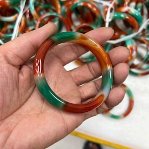 Puede incluir: Una pulsera tipo brazalete lisa y pulida con un patrón jaspeado de secciones en verde vibrante, naranja intenso y blanco translúcido. La pieza de joyería circular tiene un acabado brillante y un perfil redondeado con transiciones de color definidas.