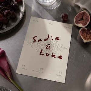 Könnte beinhalten: Eine rechteckige, cremeweiße Save-the-Date-Karte mit dunkelrotem, handgeschriebenem Text, der Sadie & Luke lautet, zentriert über einem dekorativen Punktmuster. Die Karte enthält den gedruckten Text 24 07 26, Formal Invitation To Follow.