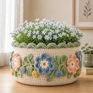 May include: A round, cream-colored crochet basket featuring a scalloped top edge and decorative floral appliqués in shades of blue, pink, and yellow with green leaves. The basket is filled with a dense arrangement of small, light blue flowers.